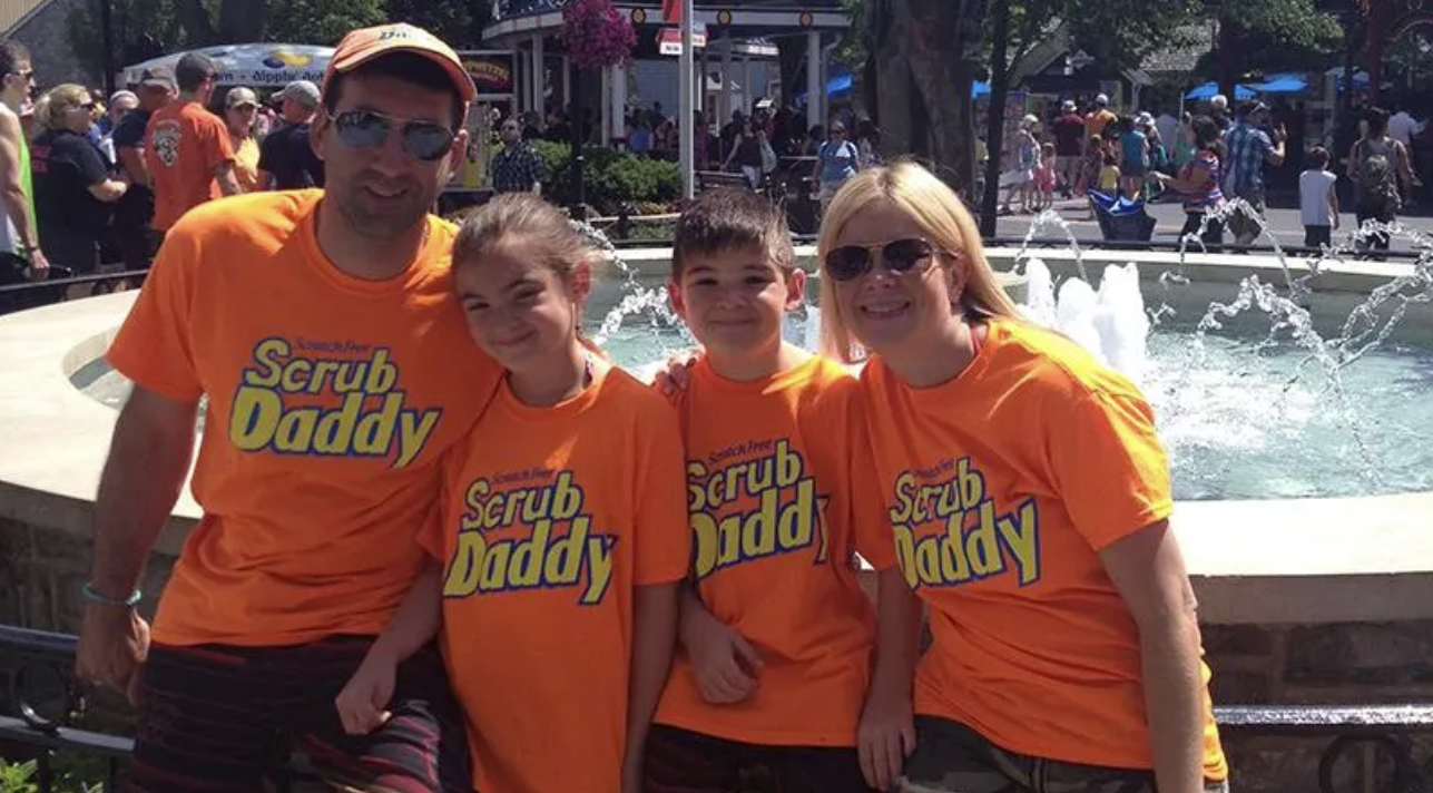 founder of scrub daddy, aaron krause posing with his children, Bryce and Sophie, and wife Stephanie Krause wearing orange scrub daddy shirts with the yellow scrub daddy logo on it. 